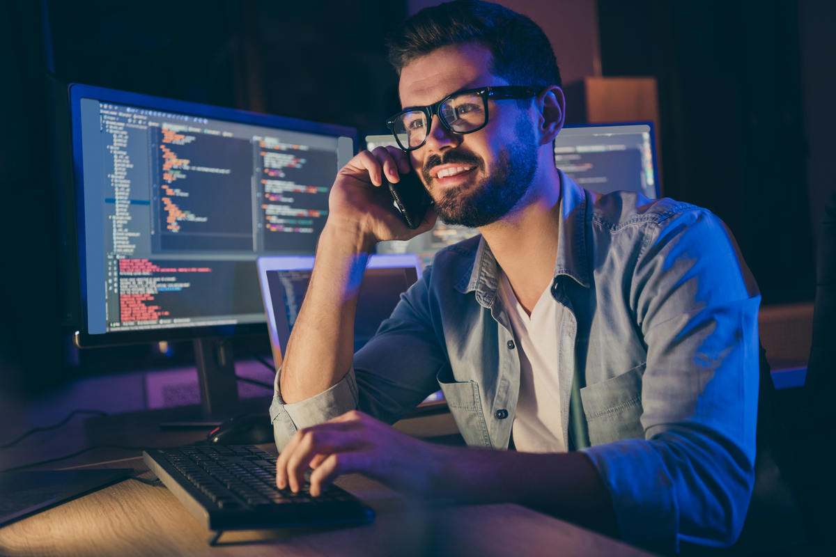 Close up portrait of his he nice attractive skilled cheerful cheery guy geek tech shark talking on phone with friend remote help assistance service in dark room workplace workstation indoors