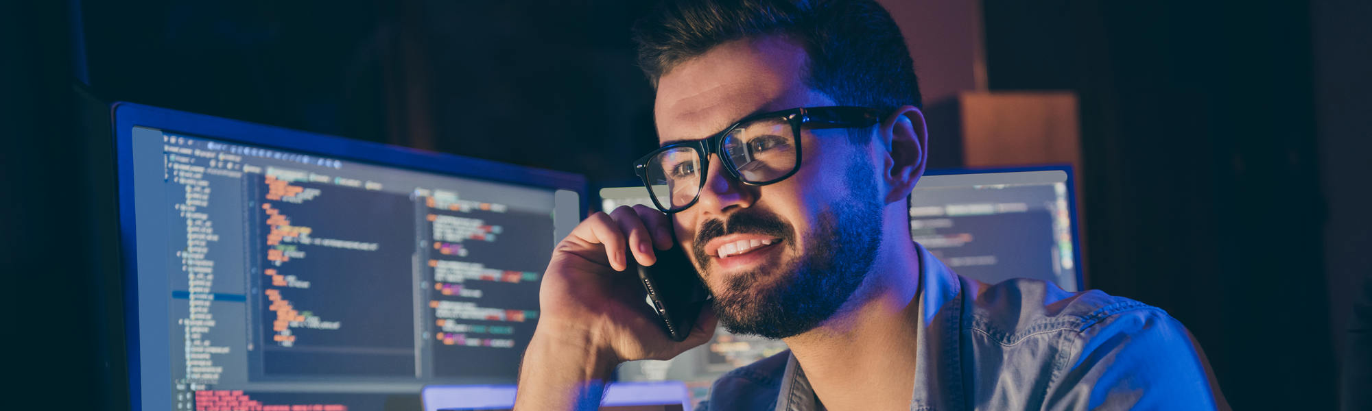 Close up portrait of his he nice attractive skilled cheerful cheery guy geek tech shark talking on phone with friend remote help assistance service in dark room workplace workstation indoors